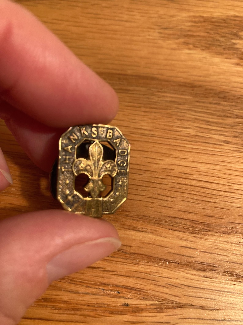 May include: A gold-colored metal pin with a fleur-de-lis design in the center. The pin is surrounded by a circular border with the words 'Thanks for Badge' inscribed around the edge.