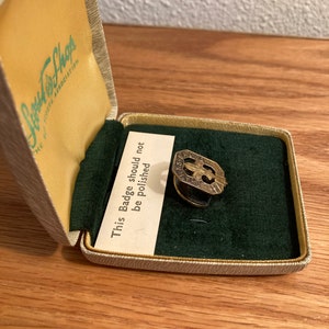 Puede incluir: Una insignia de metal de color dorado en una caja forrada de terciopelo. La caja est&aacute; etiquetada con "Junior Shops" y la insignia tiene el texto "This Badge should not be Polished".