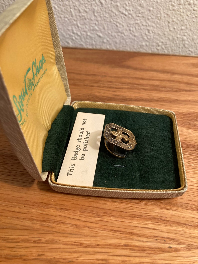 Puede incluir: Una insignia de metal de color dorado en una caja forrada de terciopelo. La caja est&aacute; etiquetada con "Junior Shops" y la insignia tiene el texto "This Badge should not be Polished".