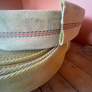 Op de afbeelding: Twee rollen brandweerslang, een is beige met een rode en zwarte streep, de andere is geel. De slang is opgerold en ligt op een houten oppervlak.