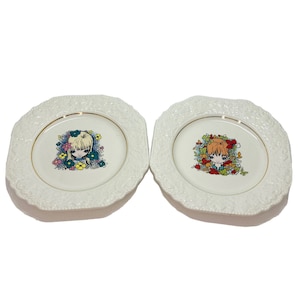 Könnte beinhalten: Zwei weiße quadratische Keramikteller mit goldener Verzierung. Jeder Teller zeigt eine farbenfrohe Illustration eines Mädchens mit Blumen. Ein Mädchen hat blonde Haare und das andere rote Haare.