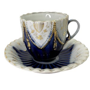 Könnte beinhalten: Ein Porzellan-Teeservice, bestehend aus einer Tasse und einer Untertasse. Die Tasse ist weiß und blau mit goldenen Akzenten und einem dekorativen Design. Die Untertasse hat einen gewellten Rand.