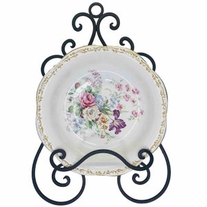 Może przedstawiać: Czarny metalowy stojak na talerze z ozdobnym wzorem w postaci zwijanych linii. Na stojaku znajduje się biały talerz ceramiczny z kwiatowym wzorem.