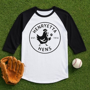 Puede incluir: Una camiseta de béisbol raglán blanca y negra con un logotipo circular que presenta una gallina sosteniendo una pelota de béisbol, con el texto "HENRYETTA HENS EST. 1914". También se ven un guante de béisbol y una pelota de béisbol.
