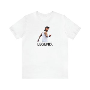 May include: White t-shirt with a person wearing a cap and a white vest, pointing a finger. The word "LEGEND." is printed in black below the image. The person has tattoos on their arms.