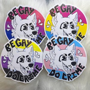 May include: Four round stickers with a white cartoon wolf illustration. Each sticker has a different LGBTQ+ pride flag background and the text "BE GAY DO CRIME". The stickers are on a white, fluffy surface.