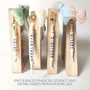 May include: Four wooden piggy bank frames with gold-tone metal clasps, each featuring a personalized beaded keychain with the names Daisy, Charlotte, Benjamin, and Amelia. Each keychain has a frayed fabric ribbon bow in pastel colors.