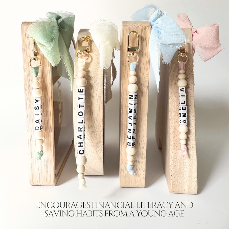 May include: Four wooden piggy bank frames with gold-tone metal clasps, each featuring a personalized beaded keychain with the names Daisy, Charlotte, Benjamin, and Amelia. Each keychain has a frayed fabric ribbon bow in pastel colors.