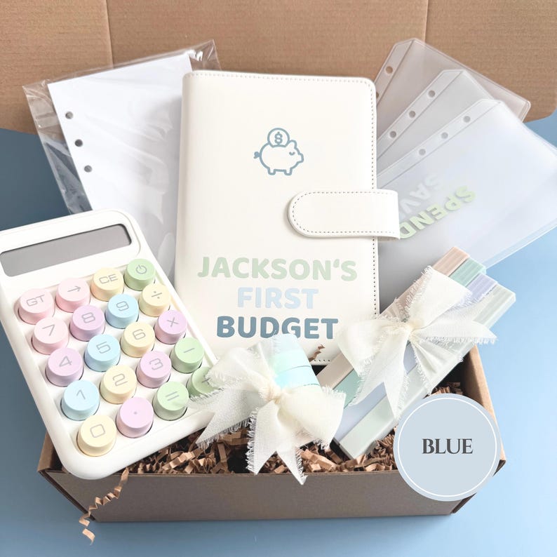 Może przedstawiać: Pudełko prezentowe zawierające biały planer budżetowy z napisem "JACKSON'S FIRST BUDGET", pastelowy kalkulator i przezroczyste etui. Pudełko zawiera r&oacute;wnież dekoracyjne wstążki i wypełniacz papierowy.