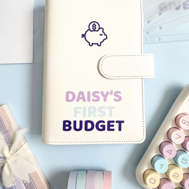 Może przedstawiać: Biały planer budżetowy z fioletową ikoną świnki skarbonki i tekstem "DAISY'S FIRST BUDGET" w kolorze fioletowym i niebieskim. Planer ma zapięcie na zatrzask i jest otoczony kalkulatorem, artykułami papierniczymi i innymi akcesoriami na jasnoniebieskiej powierzchni.