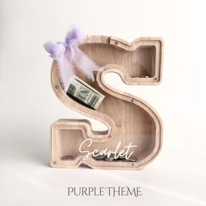Puede incluir: Una hucha de madera clara con forma de letra S y panel frontal transparente. Un lazo de color violeta claro est&aacute; atado en la parte superior izquierda. Dentro se ve un billete de un d&oacute;lar doblado y varias monedas. El nombre Scarlet est&aacute; impreso en blanco.