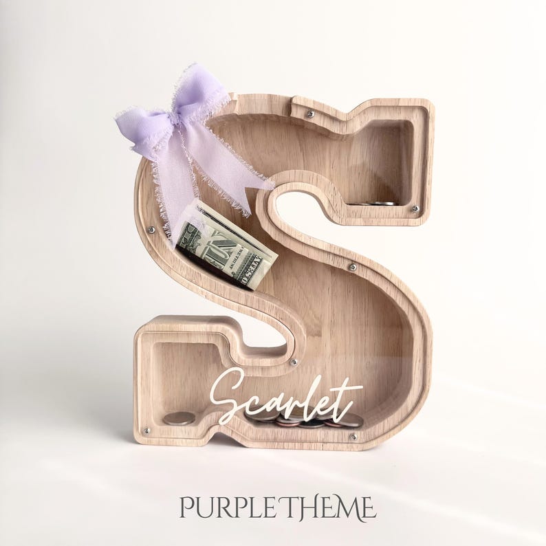 Puede incluir: Una hucha de madera clara con forma de letra S y panel frontal transparente. Un lazo de color violeta claro est&aacute; atado en la parte superior izquierda. Dentro se ve un billete de un d&oacute;lar doblado y varias monedas. El nombre Scarlet est&aacute; impreso en blanco.