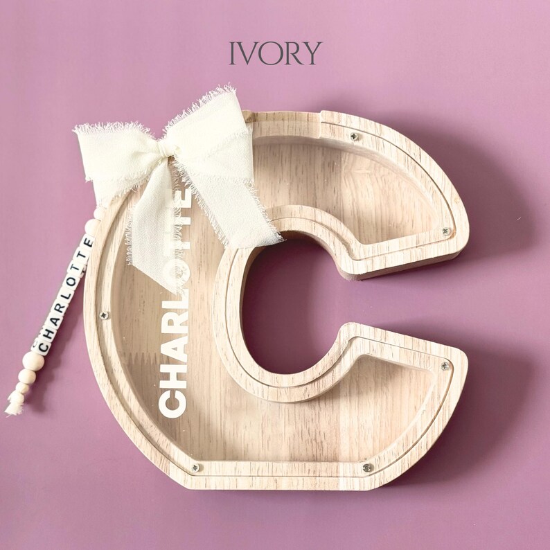 May include: A wooden letter C shaped piggy bank with a clear acrylic front panel. The name CHARLOTTE is printed in white on the acrylic. An ivory fabric bow is attached to the top left, and a string of white beads spelling CHARLOTTE hangs down.