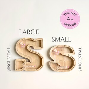 May include: Two wooden letter "S" shaped piggy banks, one large and one small, with the text "Serena" on each. The large bank is 9 inches tall, and the small bank is 7 inches tall. Both banks have decorative flowers.