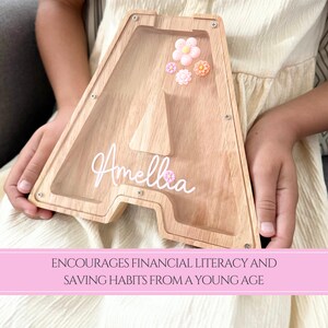 May include: Wooden letter "A" shaped piggy bank with a clear front, decorated with pink and orange flowers. The name "Amellia" is written in white script. The piggy bank is designed to encourage financial literacy.