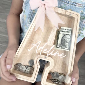 Puede incluir: Una hucha de madera de color claro con forma de letra A y un panel frontal de acr&iacute;lico transparente. El nombre Adeline est&aacute; impreso en letra cursiva blanca sobre el acr&iacute;lico. Un lazo de tela rosa p&aacute;lido est&aacute; sujeto en la parte superior y contiene monedas y un billete.