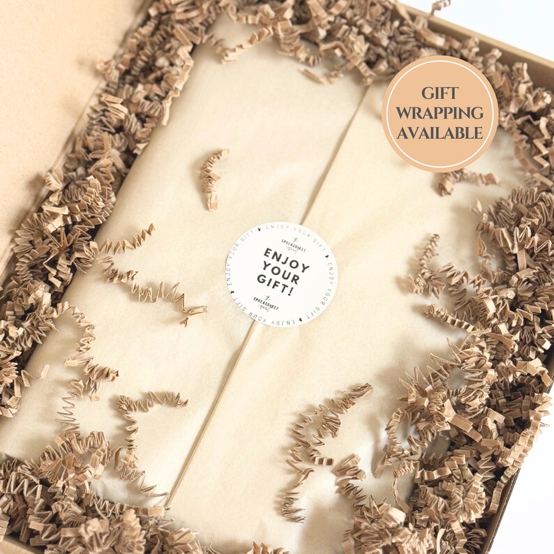 May include: A gift box with brown crinkle paper and a beige interior. The box contains a wrapped gift with a white sticker that reads "Enjoy Your Gift!". A circular sticker indicates "Gift Wrapping Available".