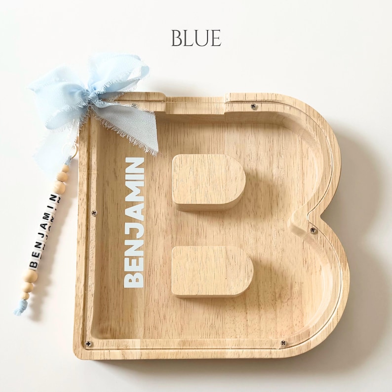 May include: A wooden letter B shaped piggy bank with a clear front panel. The name BENJAMIN is printed in white on the wood. A light blue ribbon is tied to the top left corner, attached to a string of wooden beads spelling out BENJAMIN.