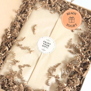 May include: A gift box with brown crinkle paper and a beige interior. The box contains a wrapped item, sealed with a round sticker that reads "Enjoy Your Gift!". An orange sticker with a gift icon and the words "Ready to Gift" is also visible.