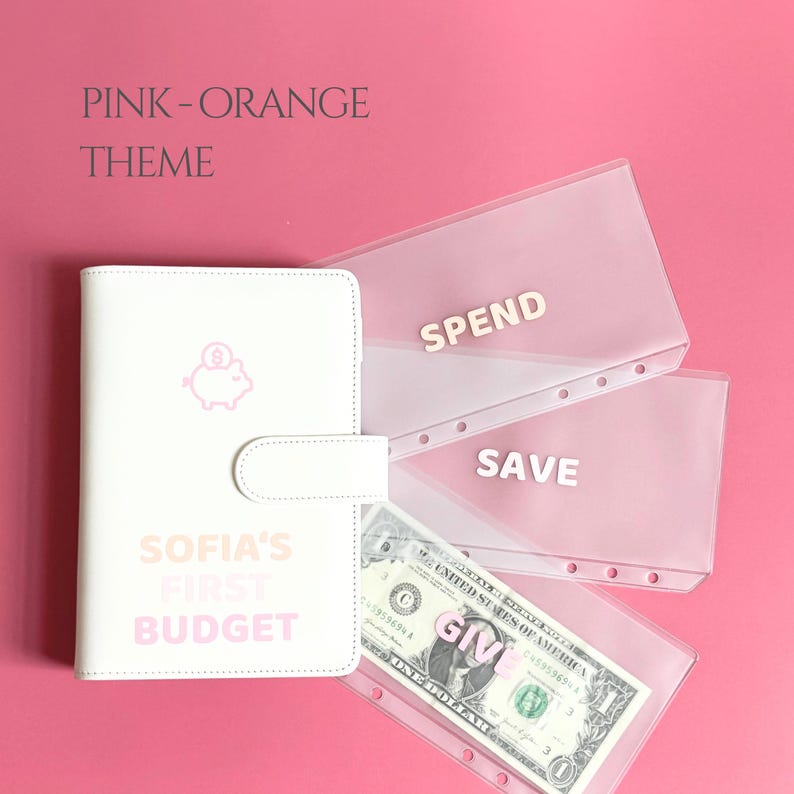May include: A white budget binder with a pink piggy bank icon and the text "SOFIA'S FIRST BUDGET." Transparent pink envelopes are labeled "SPEND," "SAVE," and "GIVE." A dollar bill is visible in the "GIVE" envelope. The theme is pink and orange.