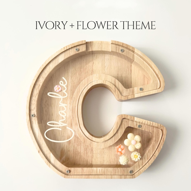May include: Wooden letter "C" shaped bank with a clear front. The bank has the name "Charlie" in cursive script and is decorated with small flower embellishments. The text "IVORY + FLOWER THEME" is at the top.