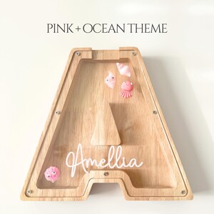 May include: Wooden letter "A" shaped bank with a clear front, decorated with pink ocean-themed embellishments. The bank has the name "Amellia" in white script. The text "PINK + OCEAN THEME" is at the top.