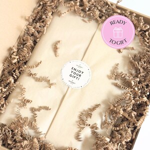 May include: A brown cardboard gift box filled with crinkled brown paper filler. The center contains a package wrapped in light beige tissue paper, sealed with a round white sticker that reads ENJOY YOUR GIFT. A pink circular badge says READY TO GIFT.