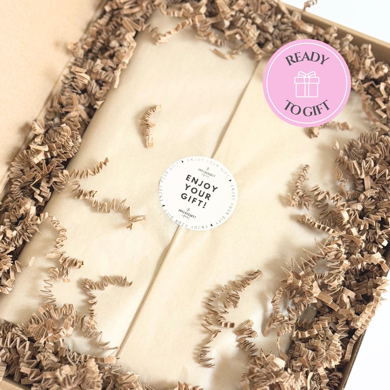 May include: A brown cardboard gift box filled with crinkled brown paper filler. The center contains a package wrapped in light beige tissue paper, sealed with a round white sticker that reads ENJOY YOUR GIFT. A pink circular badge says READY TO GIFT.