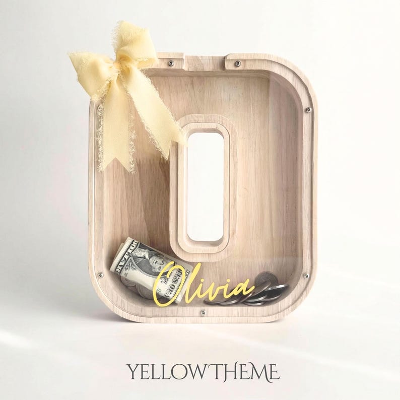 Puede incluir: Una hucha de madera con forma de letra O y panel frontal transparente, con un lazo de tela amarillo claro en la esquina superior izquierda. El nombre Olivia est&aacute; escrito en amarillo en la parte frontal, con un billete enrollado y monedas dentro.