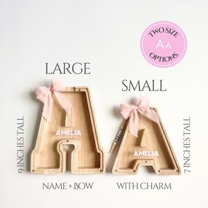 May include: Two wooden letter A piggy banks labeled LARGE and SMALL. The large bank is 9 inches tall with a pink bow. The small bank is 7 inches tall with a pink bow and a name charm. Both feature the name AMELIA printed in white on the front.