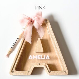 May include: A wooden letter A shaped piggy bank with a clear front panel. The name AMELIA is printed in white on the wood. A light pink fabric bow is attached to the top, and a string of wooden beads with the name AMELIA hangs from the side.
