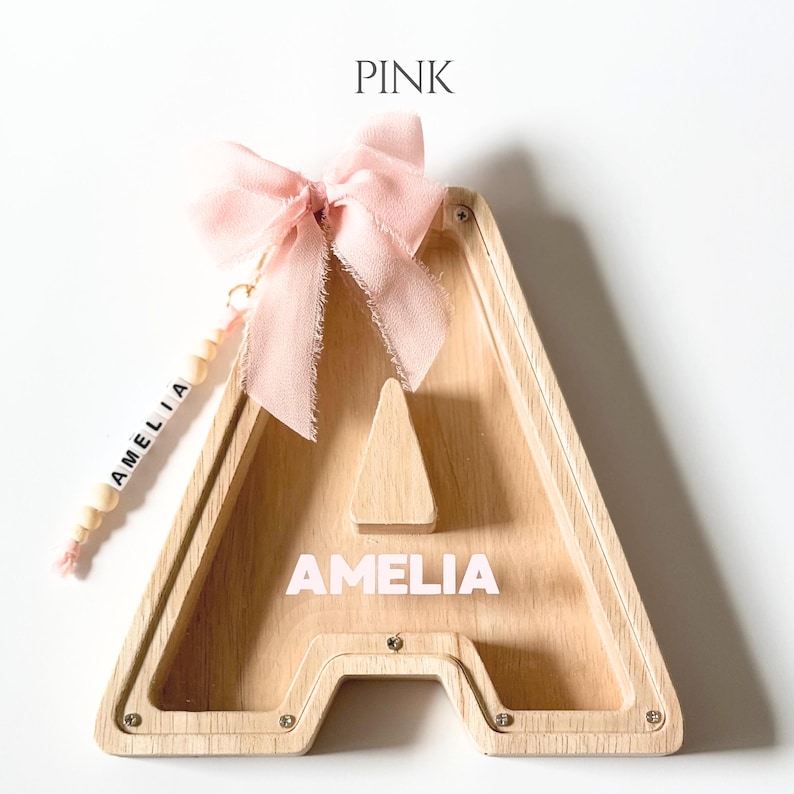May include: A wooden letter A shaped piggy bank with a clear front panel. The name AMELIA is printed in white on the wood. A light pink fabric bow is attached to the top, and a string of wooden beads with the name AMELIA hangs from the side.