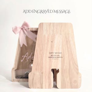 Puede incluir: Una hucha de madera clara con forma de letra A, panel frontal transparente, un lazo rosa en la parte superior y un mensaje grabado que dice "HAPPY 1ST BIRTHDAY! WE LOVE YOU, GRANDMA & GRANDPA" junto al nombre "Adele".