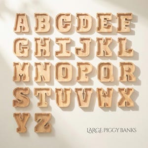 Puede incluir: Una colecci&oacute;n de veintis&eacute;is huchas de madera de color claro con forma de letra, dispuestas en orden alfab&eacute;tico de la A a la Z. Cada letra tiene un centro hueco y un panel frontal transparente. El texto LARGE PIGGY BANKS aparece abajo a la derecha.