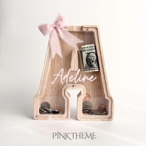 Puede incluir: Una hucha de madera clara con forma de letra A y un panel frontal de acr&iacute;lico transparente. El nombre Adeline est&aacute; impreso en letra cursiva blanca en la parte delantera. Un lazo de tela rosa suave est&aacute; sujeto en la parte superior y el interior contiene monedas y un billete.