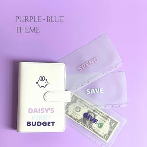 May include: A white budget planner with the text "DAISY'S FIRST BUDGET" and a piggy bank icon. Includes three transparent pouches labeled "SPEND", "SAVE", and "GIVE", with a one-dollar bill visible. The theme is purple and blue.