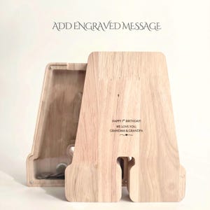 May include: A light-colored wooden piggy bank shaped like the letter A with a clear acrylic front panel. The wood features an engraved message that reads: HAPPY 1ST BIRTHDAY! WE LOVE YOU, GRANDMA & GRANDPA. The text ADD ENGRAVED MESSAGE appears above.