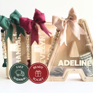 May include: Wooden letter-shaped money banks with clear fronts. Each bank has a name on it, adorned with a ribbon bow and a beaded name tag. One bank contains coins and a dollar bill. The image also includes two round stickers.