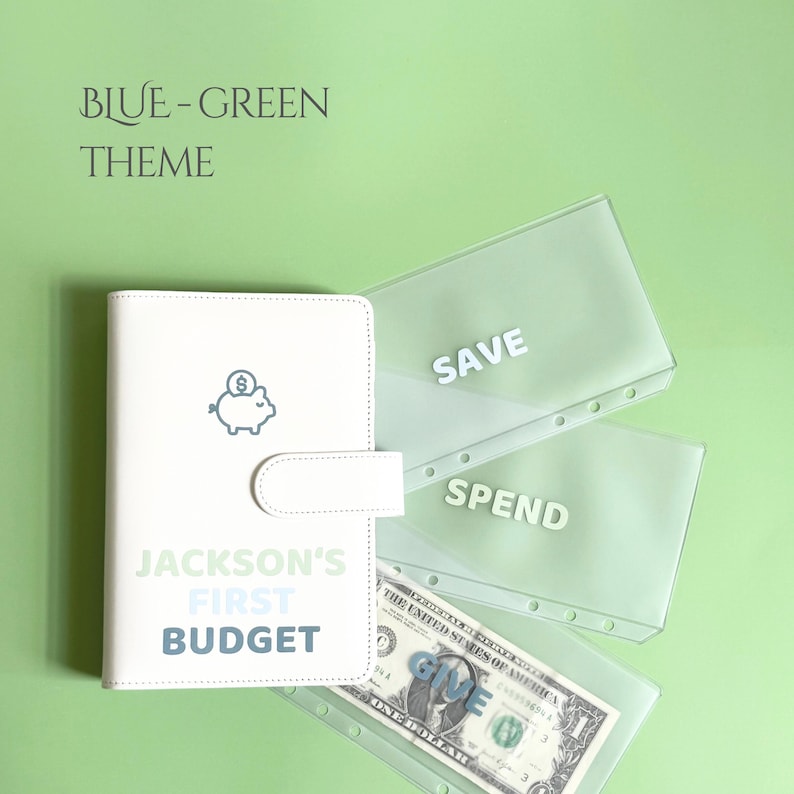 May include: A white budget binder with a piggy bank icon and the text "JACKSON'S FIRST BUDGET" in blue and green. Includes clear plastic envelopes labeled "SAVE" and "SPEND", and a dollar bill. The theme is blue-green.