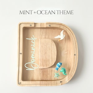 May include: Wooden piggy bank shaped like the letter "D" with the name "Dominick" in mint green script. The bank is decorated with a white dove, a blue fish, a green seahorse, and a starfish. The text "MINT + OCEAN THEME" is at the top.