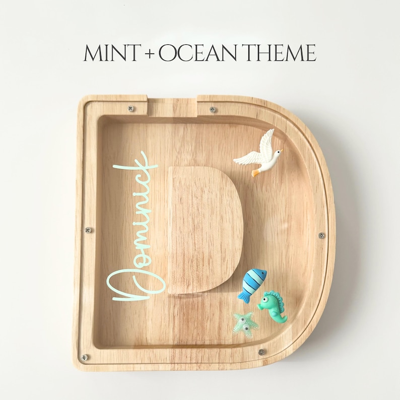 May include: Wooden piggy bank shaped like the letter "D" with the name "Dominick" in mint green script. The bank is decorated with a white dove, a blue fish, a green seahorse, and a starfish. The text "MINT + OCEAN THEME" is at the top.