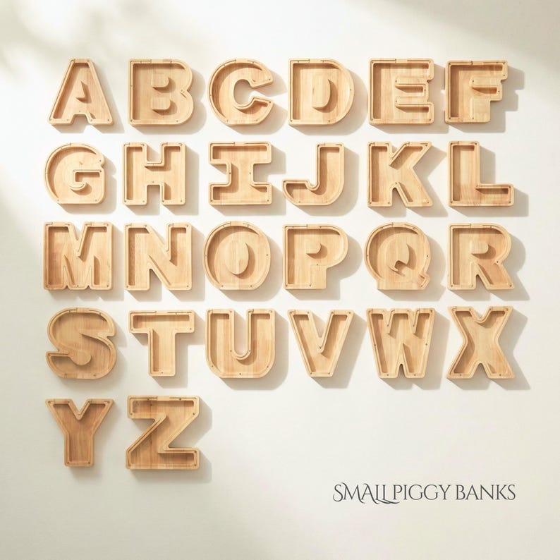Puede incluir: Una colecci&oacute;n de veintis&eacute;is huchas de madera de color claro con forma de letra, cada una con un panel frontal transparente. Las letras de la A a la Z est&aacute;n dispuestas en filas, con el texto SMALL PIGGY BANKS impreso en la esquina inferior derecha.