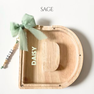 May include: A wooden letter D-shaped piggy bank with a clear front panel. The name DAISY is printed in light green on the wood. A sage green fabric bow is attached to the top left, featuring a dangling charm with wooden beads and the name DAISY.