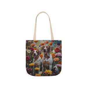 Puede incluir: Bolso tote crema con una imagen colorida de dos perros en un campo de flores. Los perros tienen pelaje manchado marrón y blanco y llevan collares. El bolso tiene un asa beige. Las flores son rojas, naranjas y amarillas.