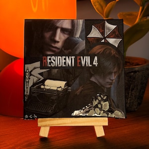 May include: A square canvas print featuring a collage of Resident Evil 4 imagery, including the game title, a character portrait, a typewriter, and the Umbrella Corporation logo. The artwork is displayed on a small wooden easel stand.