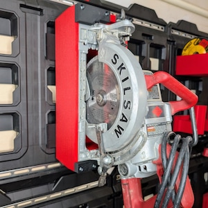 May include: A silver and red SKILSAW circular saw mounted on a black and red Milwaukee Packout storage system. The saw blade is visible, and the handle is red. The storage system has multiple compartments.