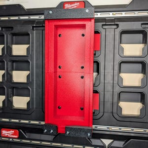 May include: A red and black Milwaukee Packout mounting system. The red bracket is mounted on a black storage unit. The Milwaukee logo is visible at the top and bottom of the image. The bracket is secured with black screws and foam padding.