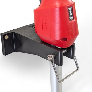 May include: A black bracket with a red power tool attached. The bracket is mounted on a silver pole. The power tool has the brand name "M18" printed on it.