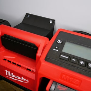 Peut inclure: Un chargeur d'outils électriques Milwaukee M18 Fuel rouge monté sur un support mural noir avec deux vis. Le chargeur dispose d'un écran numérique et de boutons pour contrôler le processus de charge.