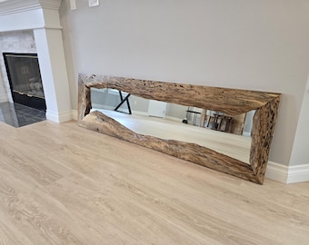 May include: A long, rectangular mirror featuring a thick, natural-edge wooden frame. The frame displays a rough, textured surface with visible knots, grain patterns, and a medium-brown finish, surrounding a clear, reflective glass surface.